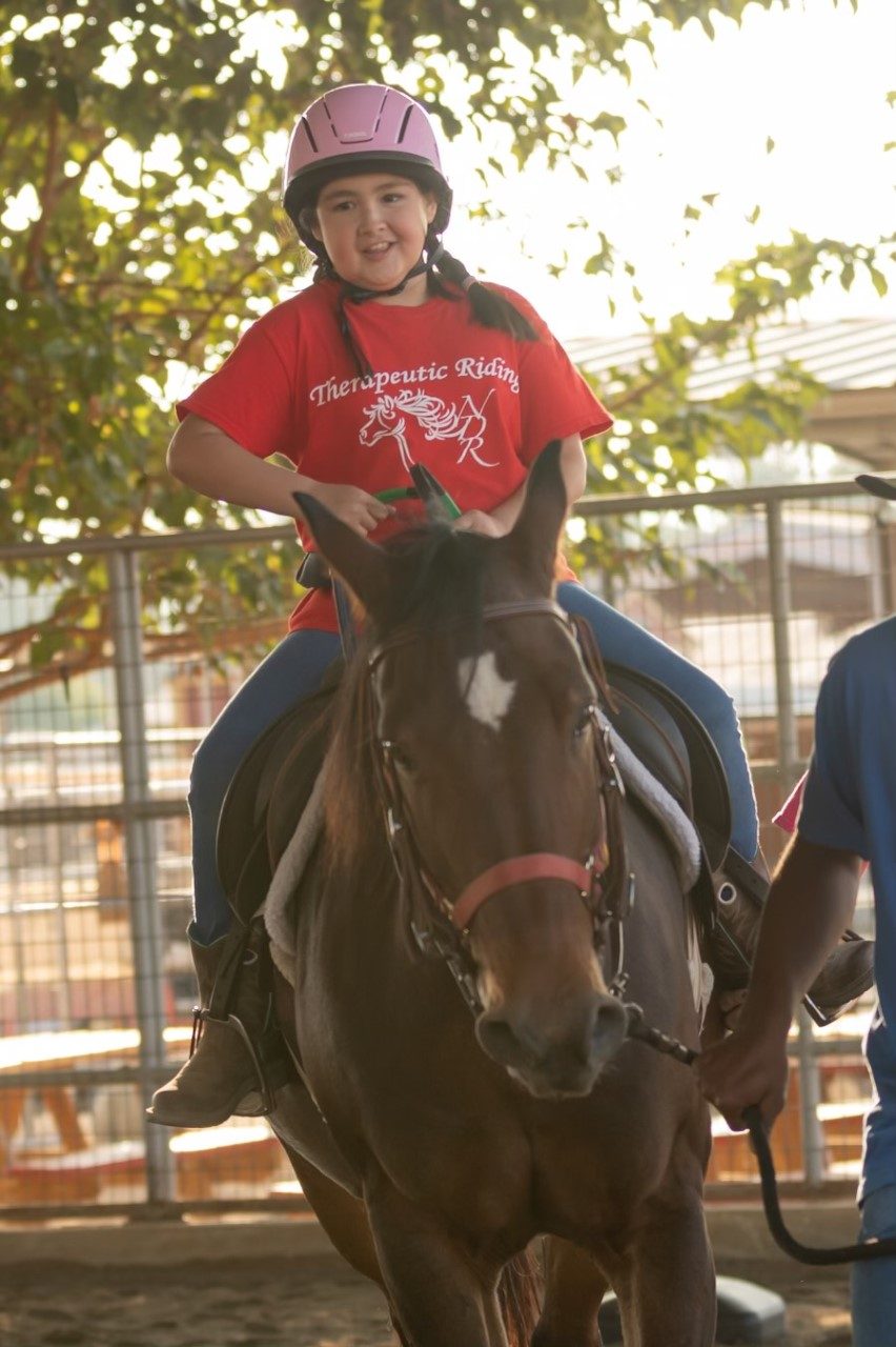 A smiling rider in a pink helmet riding a horse through the arena